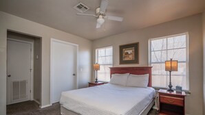 Une chambre à coucher avec un lit, deux tables de chevet, un ventilateur de plafond et une fenêtre avec des stores.