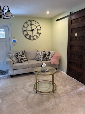 Una sala de estar con un sofá color beige, cojines estampados, una mesa de centro redonda de vidrio y un reloj de pared grande.