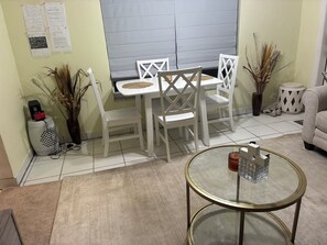 A dining area with a white table and chairs, a small round table with a glass top, and a decorative vase with dried plants.