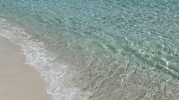 Una playa con agua turquesa cristalina, una orilla arenosa y un muelle a lo lejos.