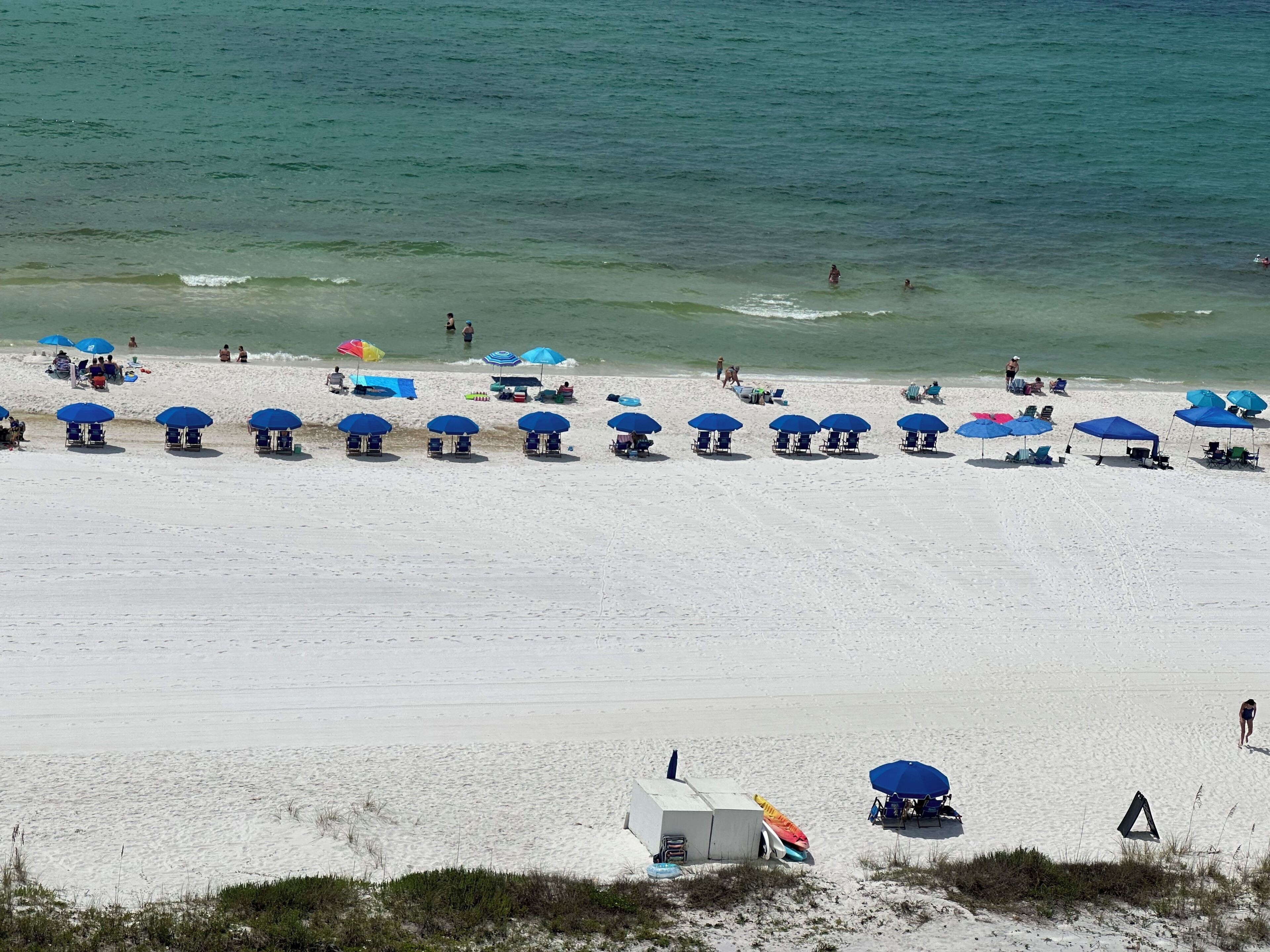 Ein Strand mit blauen Sonnenschirmen und Liegestühlen, helles Sand und das Meer im Hintergrund.