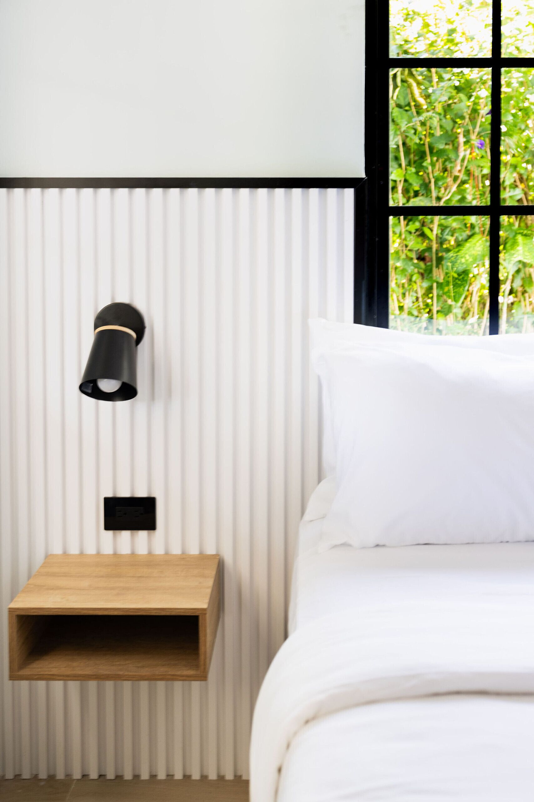 Een slaapkamer met een bed, nachtkastje, wandlamp en een raam met uitzicht op groen.