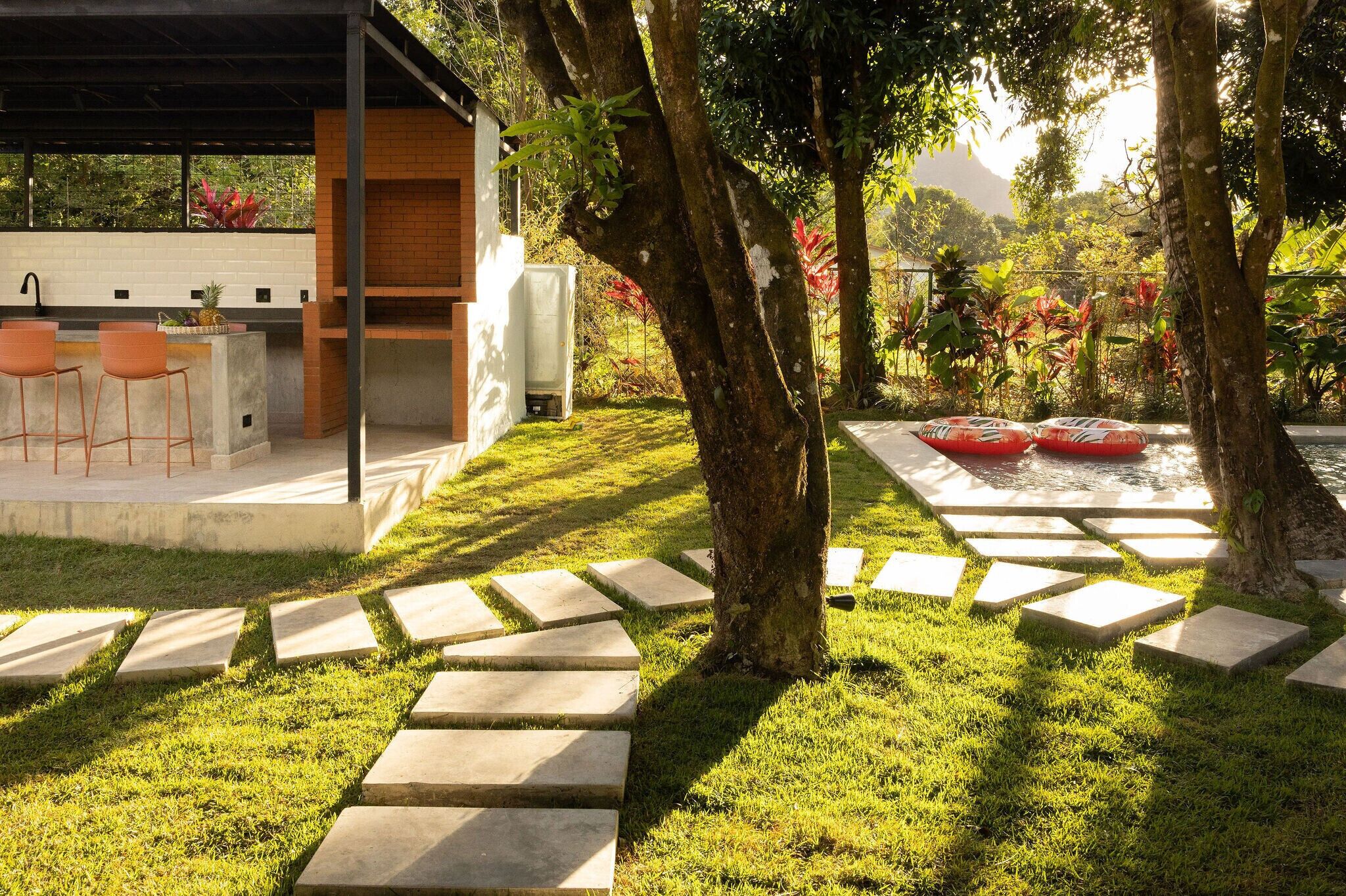 Un área de cocina exterior moderna con barra y asientos, rodeada de exuberante vegetación y un camino de piedra.