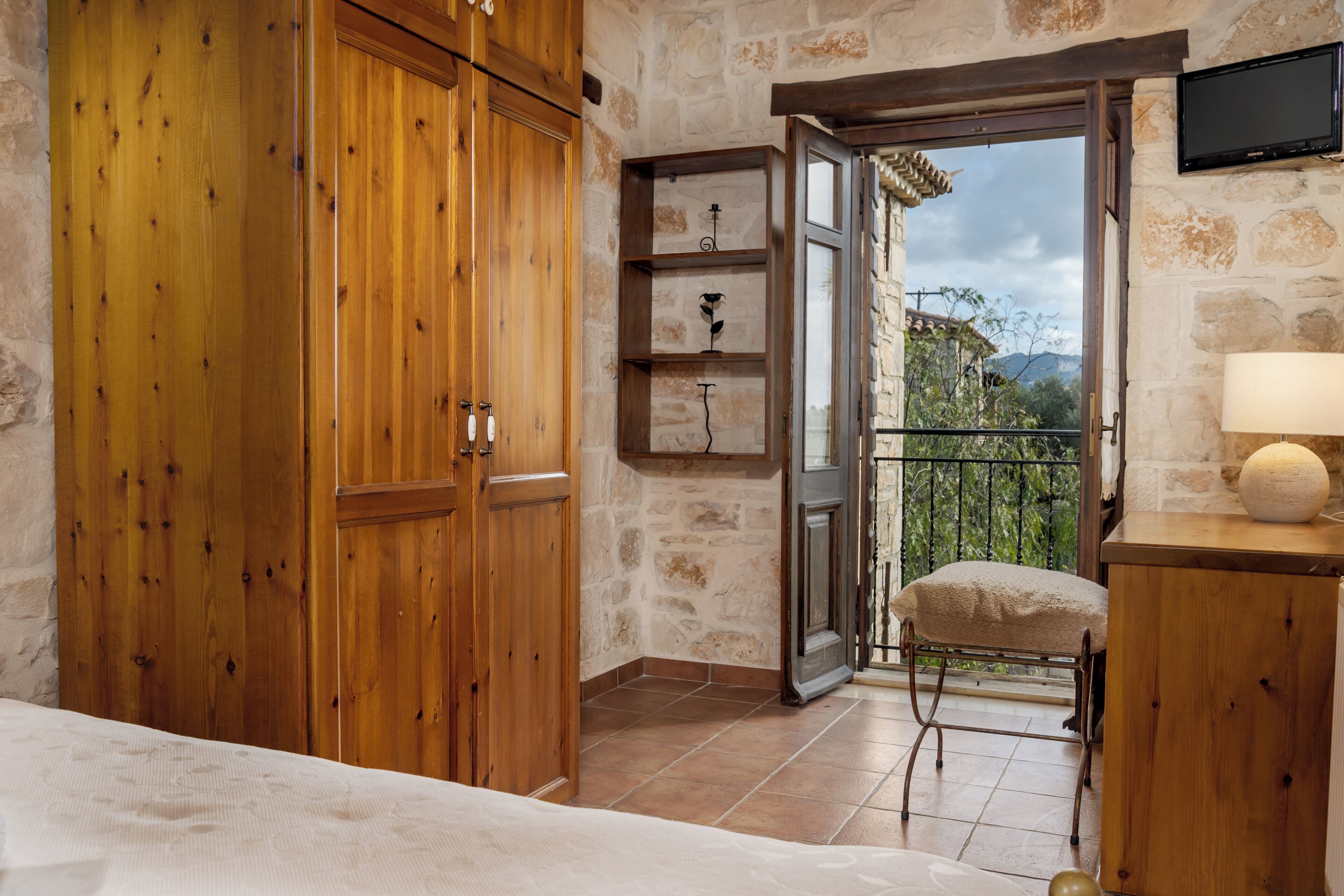 Un dormitorio con armarios de madera, una mesita de noche con lámpara, un taburete pequeño y vistas al exterior a través de una puerta abierta.