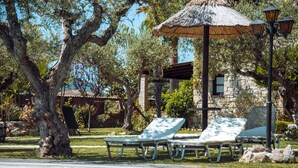 A pool area with lounge chairs, a thatched umbrella, and a stone building in the background. - Grantzaos Villas Zante (Agios Sostis)