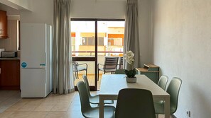 Une cuisine compacte équipée d’un réfrigérateur, d’une table à manger avec des chaises, et donnant par une porte coulissante sur un espace extérieur aménagé avec des chaises.