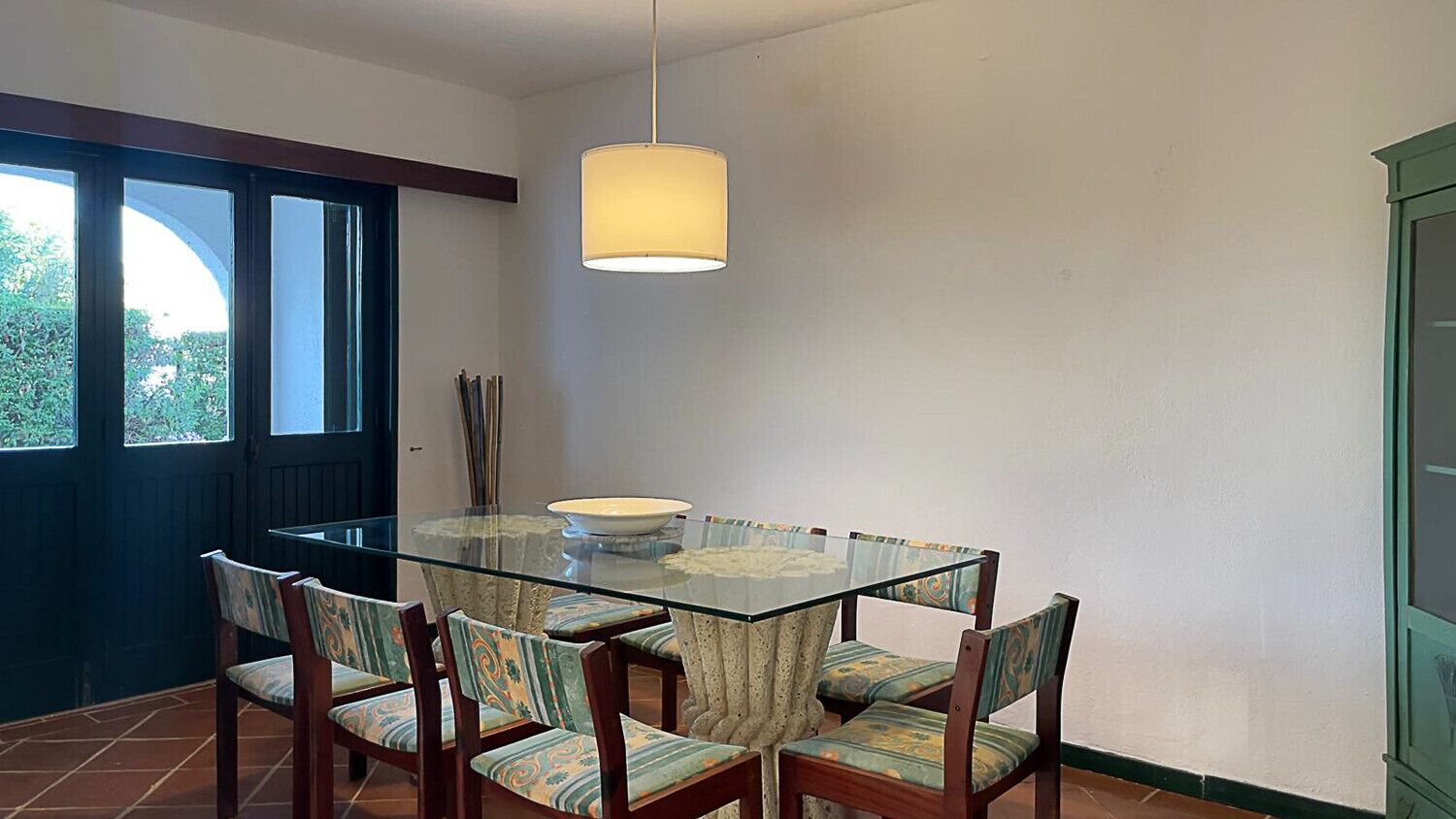 A dining area with a glass table and chairs, a hanging light fixture, and a view through a window.