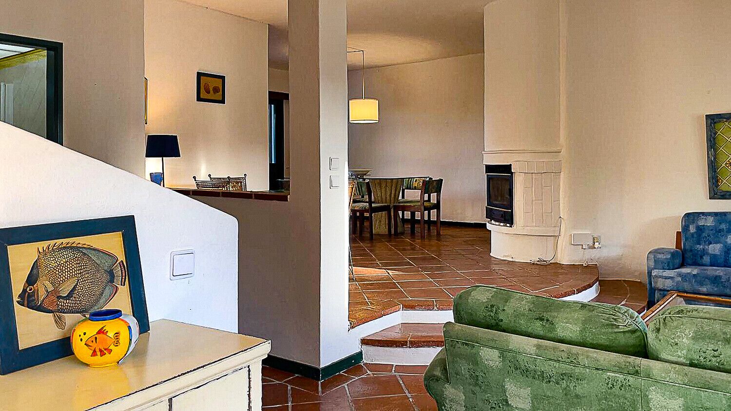 Una sala de estar con un sofá verde, un comedor con una mesa y sillas, una chimenea y una escalera que sube a otro nivel.