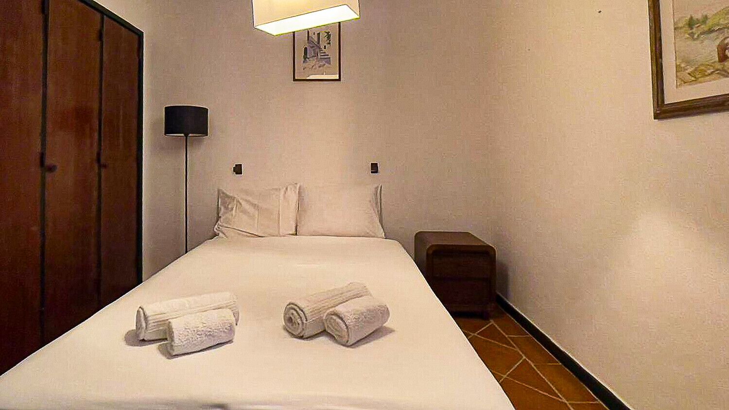 A hotel room with a bed, two rolled towels, a bedside lamp, a small bedside table, and a picture on the wall.