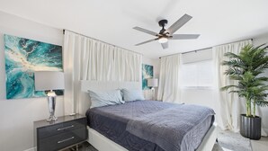 Une chambre à coucher comprenant un lit, une table de chevet, un ventilateur de plafond, une grande fenêtre avec des rideaux et une plante en pot.