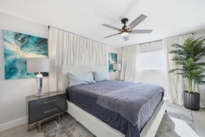A bedroom with a bed, a nightstand, a ceiling fan, a large window with curtains, and a potted plant. - 5 Min to Madeira Beach • Private Pool • Games (Seminole)