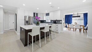 Une cuisine moderne avec des meubles blancs, un îlot central doté de tabourets, un coin repas avec une table et des chaises, et une vue sur l’extérieur grâce à de grandes fenêtres.