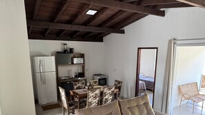 Una sala de estar con sofá color beige, una zona de comedor con mesa y sillas, una cocina con electrodomésticos y un techo alto con vigas de madera.