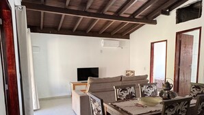 Amplia sala de estar con techo alto y vigas de madera a la vista. Incluye una mesa de comedor con sillas, un sofá y un televisor al fondo.