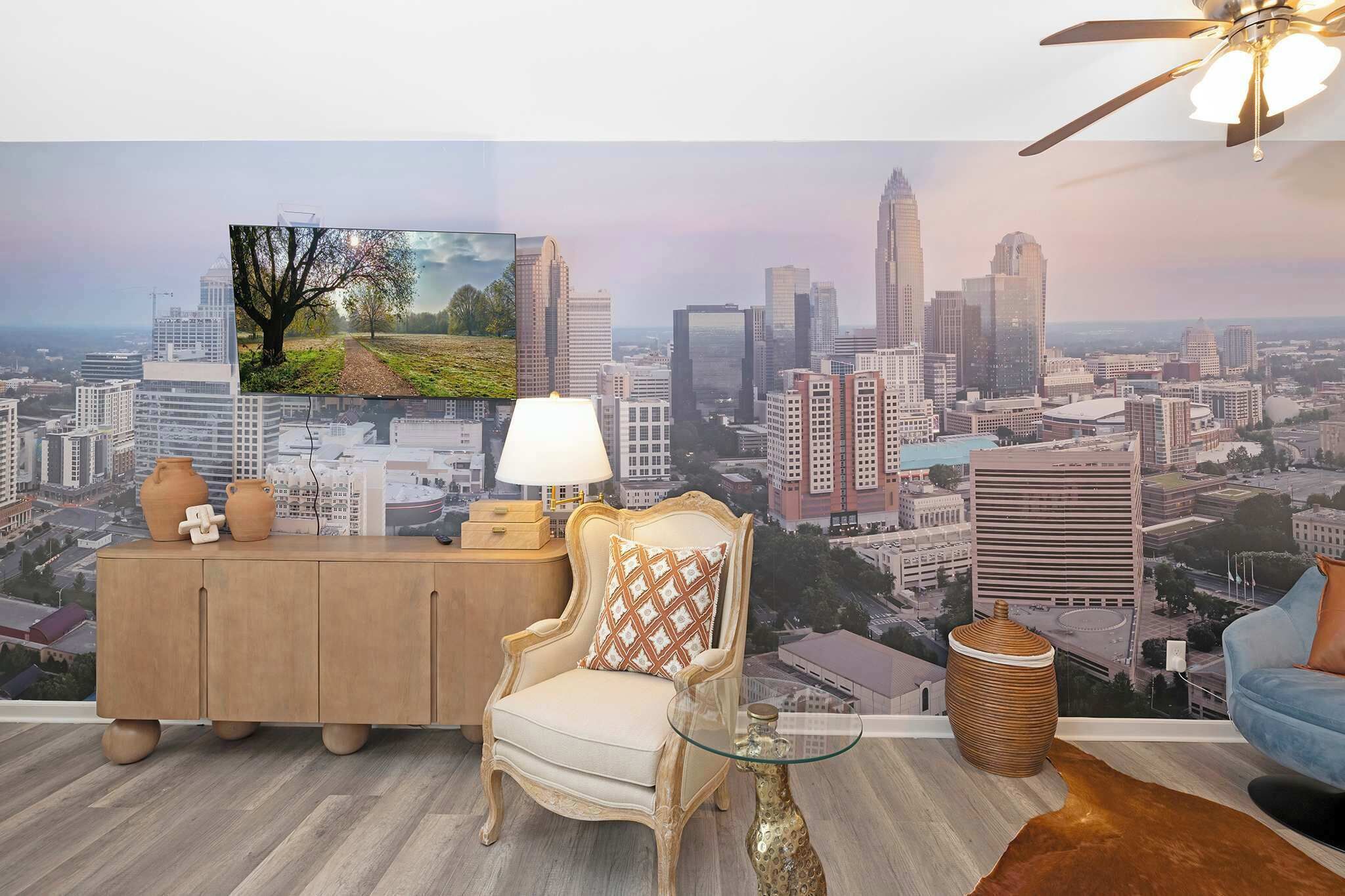A room with a cityscape mural, a wooden cabinet with decorative items, a chair, a table with a lamp, and a ceiling fan.