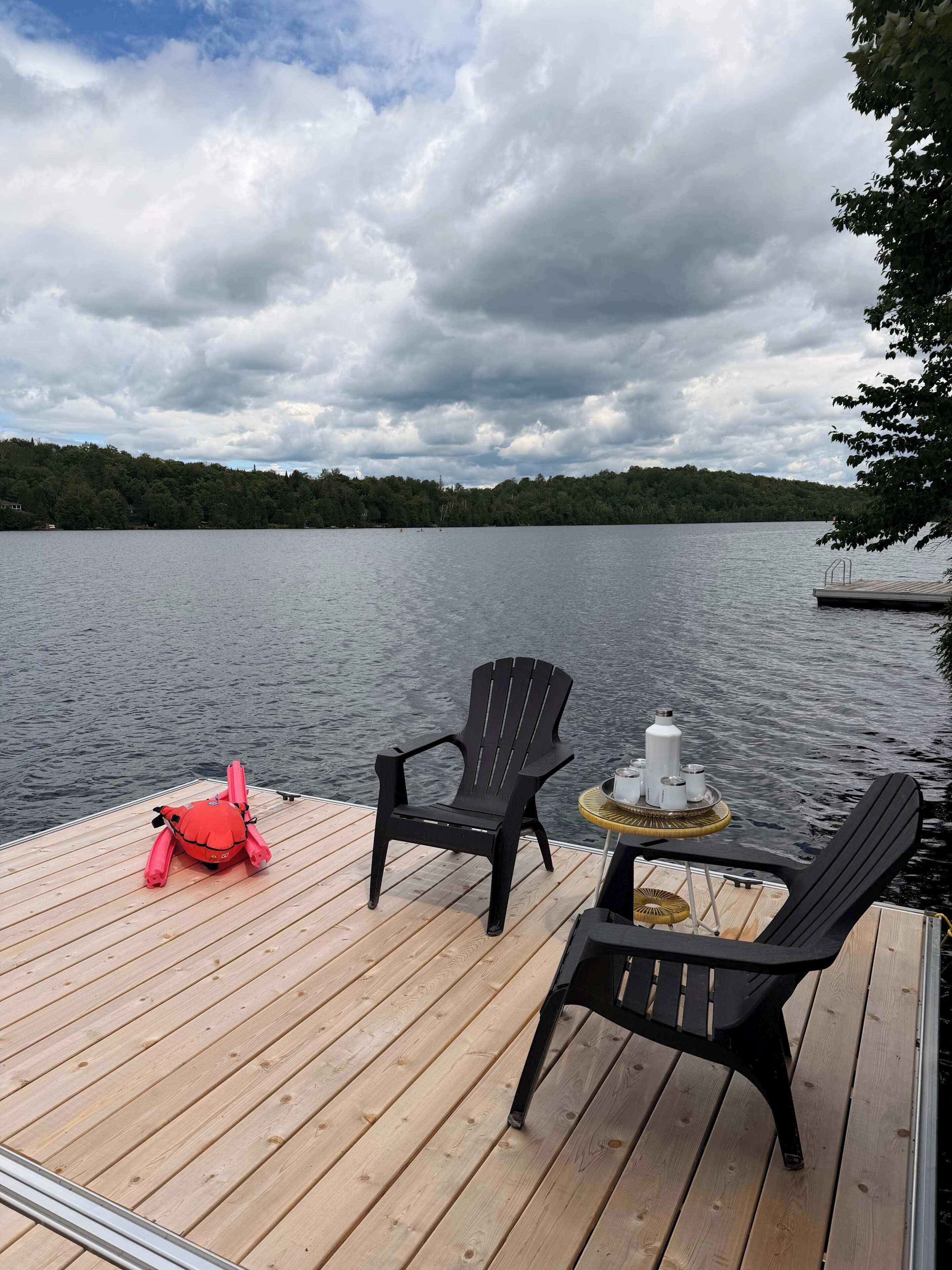 Eine Holzterrasse mit zwei schwarzen Stühlen und einem roten Kissen in Form eines Krebsses, ein kleiner runder Tisch mit einer weißen Flasche und einem Korb, sowie der Blick auf einen See mit bewaldeter Uferlinie unter einem bewölkten Himmel.