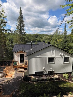 A cabin with a deck, a chimney, and a view of a forested area. - Waterfront & Spa | Chalet Lac Clair (Sainte-Marguerite-du-Lac-Masson)