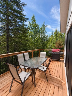 A deck with a table and chairs, a grill, and a view of trees. - Waterfront & Spa | Chalet Lac Clair (Sainte-Marguerite-du-Lac-Masson)