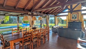 A covered outdoor dining area with a wooden table and chairs, a grill, and a view of a pool and greenery.