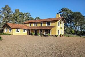 A yellow two-story house with a red-tiled roof, a covered porch, and a large gravel driveway. - Gorgeous 7-bedroom farmhouse with AC in charming Soca (Departamento de Canelones)