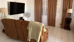 A living room with a brown sofa, a flat-screen TV, a standing fan, and a window with brown curtains. - Cozy 3-bedroom house in serene Carolina with AC (Carolina)