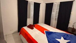 A bedroom with a bed covered in a flag-themed bedspread, black curtains, and an air conditioning unit on the wall. - Cozy 3-bedroom house in serene Carolina with AC (Carolina)