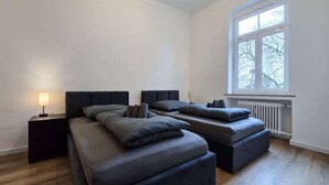 A bedroom with two beds, a nightstand, a window, and a radiator. - Alpha Apartments: 2BR for 4, Quiet Retreat (Krefeld)