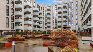 Un immeuble résidentiel à plusieurs étages, doté de balcons, d’une terrasse en bois et d’espaces aménagés avec des arbustes et des arbres.