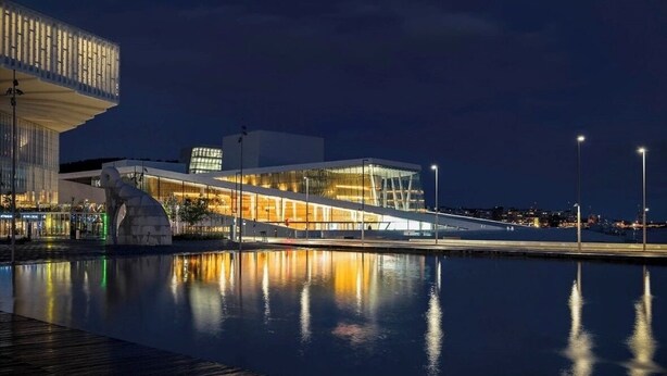Un bâtiment moderne doté d’une façade en verre et d’une rampe menant à l’entrée, situé au bord d’un plan d’eau paisible qui reflète les lumières.