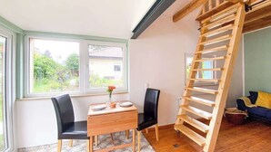 A room with a wooden table and chairs, a wooden ladder, and large windows with a view of greenery. - Tiny house in Rangsdorf for 4 people + fireplace (Rangsdorf)