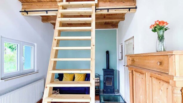 A loft bedroom with a wooden ladder leading to a sloped ceiling, a built-in wardrobe, and a wood-burning stove. - Tiny house in Rangsdorf for 4 people + fireplace (Rangsdorf)