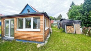 A wooden cabin with a blue roof, a glass door, and a small shed with a pile of firewood. - Tiny house in Rangsdorf for 4 people + fireplace (Rangsdorf)