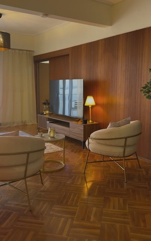 Una sala de estar moderna con suelo de madera en espiga, un televisor de pantalla plana grande, un sillón cómodo, una mesita de centro redonda y una lámpara.