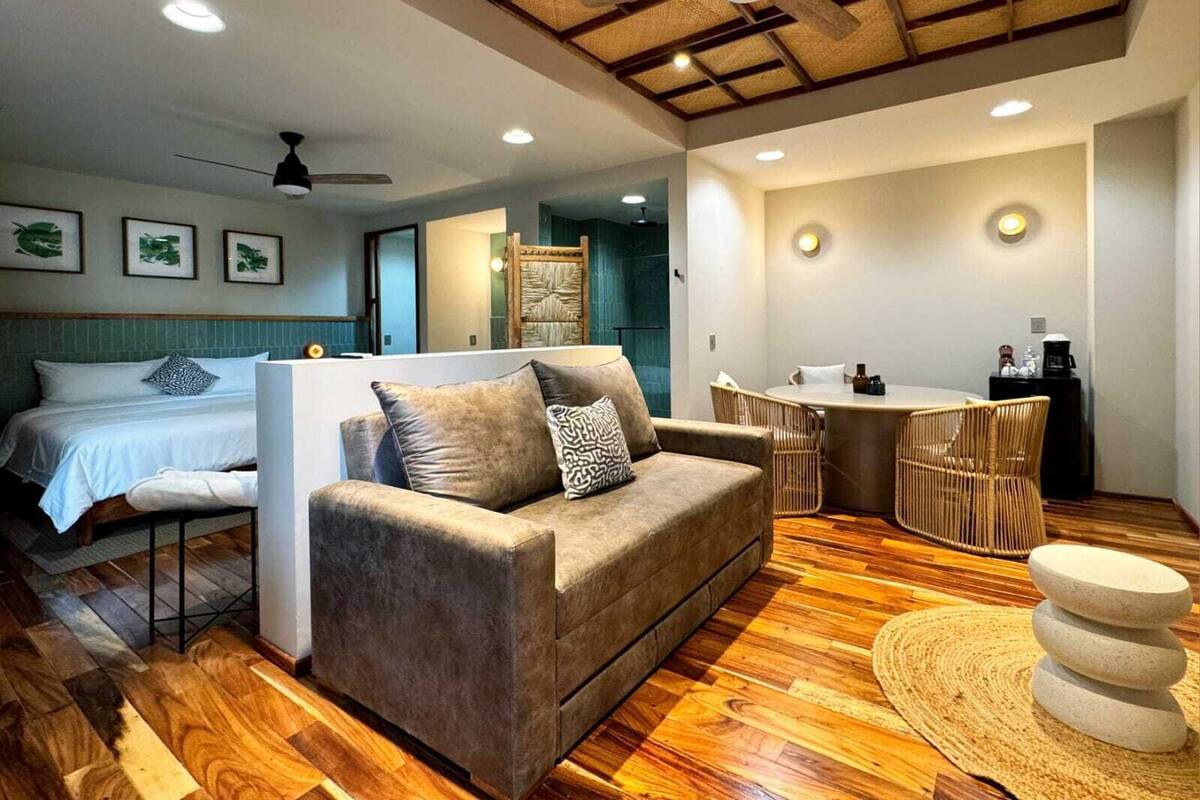 Una habitación de hotel moderna con piso de madera, un sofá, una cama, un comedor y un ventilador de techo.