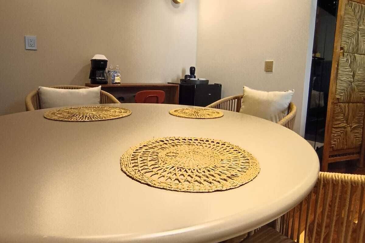 Una mesa redonda para comer con individuales trenzados, sillas de mimbre y una máquina de café sobre una encimera.