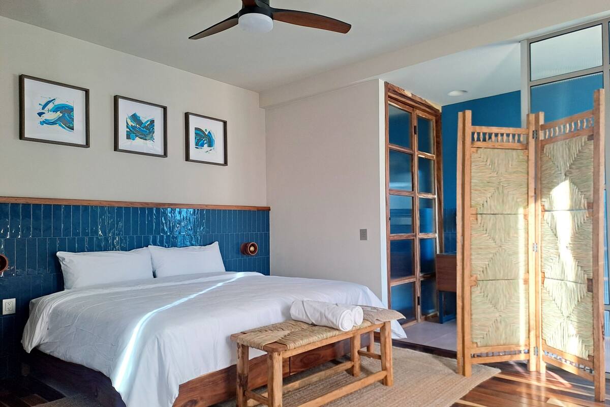 Un dormitorio con una cama, un banco de madera, un ventilador de techo y tres cuadros en la pared.