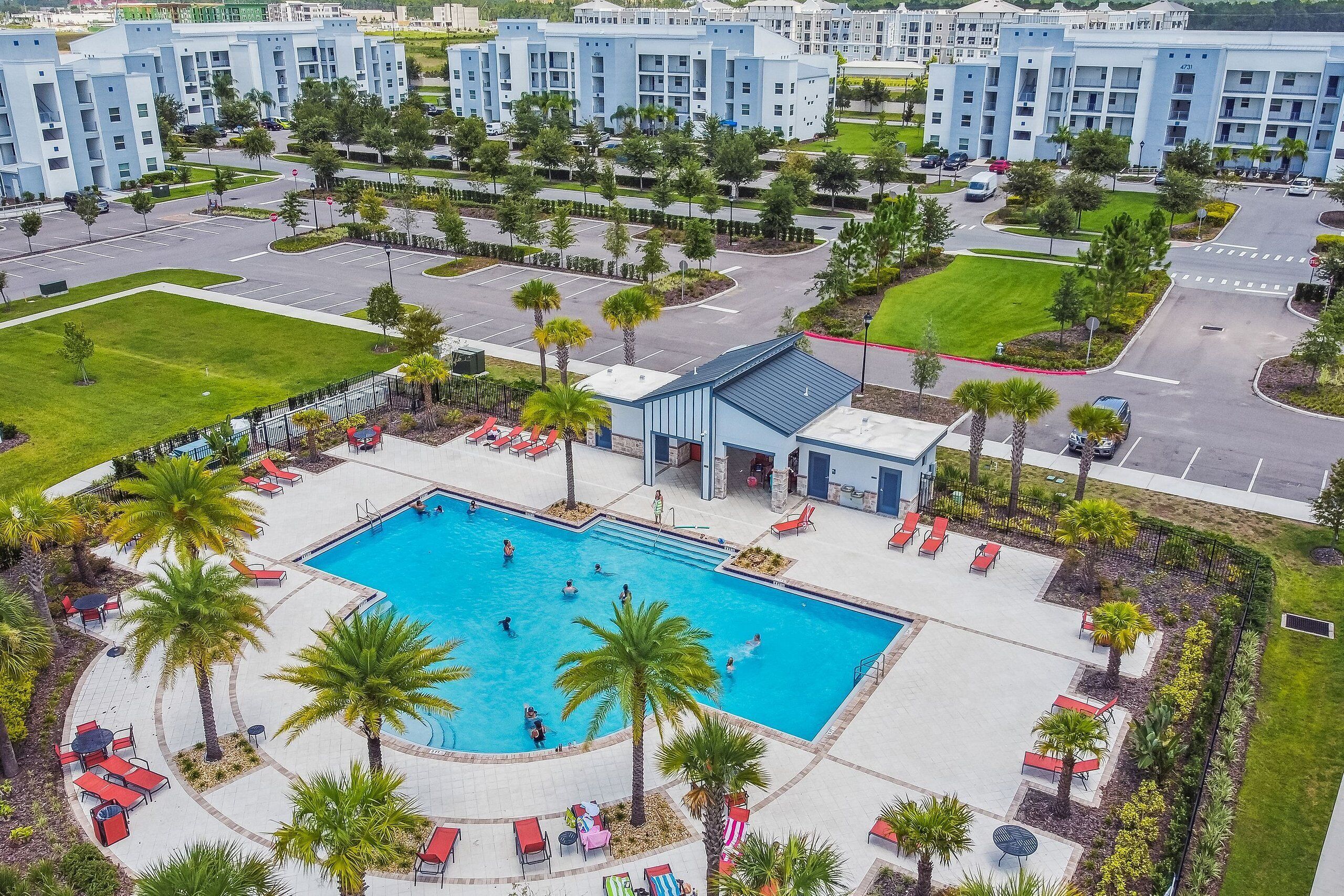A large swimming pool surrounded by palm trees and red lounge chairs, with a modern building complex in the background.
