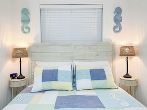 A bedroom with a wooden headboard, two bedside lamps, a bed with a patchwork quilt, and decorative seahorse wall art. - Charming 1 BD with Ocean Views/Pool/Quiet North End Complex (Tybee Island)