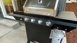 Una parrilla NEXGRILL con cuatro quemadores, una pantalla digital y un quemador lateral, ubicada en un patio de concreto frente a una casa.