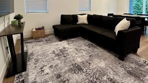 Una sala de estar moderna con un sofá seccional negro, dos ventanas con persianas, un tapete gris con estampado, una mesita con una planta y un área de comedor al fondo.