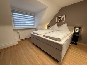 Una camera da letto con un letto, una finestra con persiane, un termosifone, un comodino con una lampada e un'opera d'arte appesa al muro.