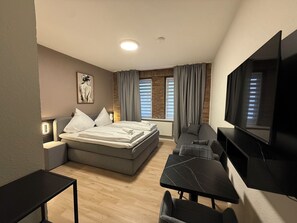 Una moderna camera d'albergo con un letto, una scrivania, una sedia, una TV e un'ampia finestra con tende.
