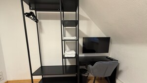 A compact room with a black metal desk, a grey chair, a TV, and a built-in shelving unit. - SHIN CENTRAL (Essen)