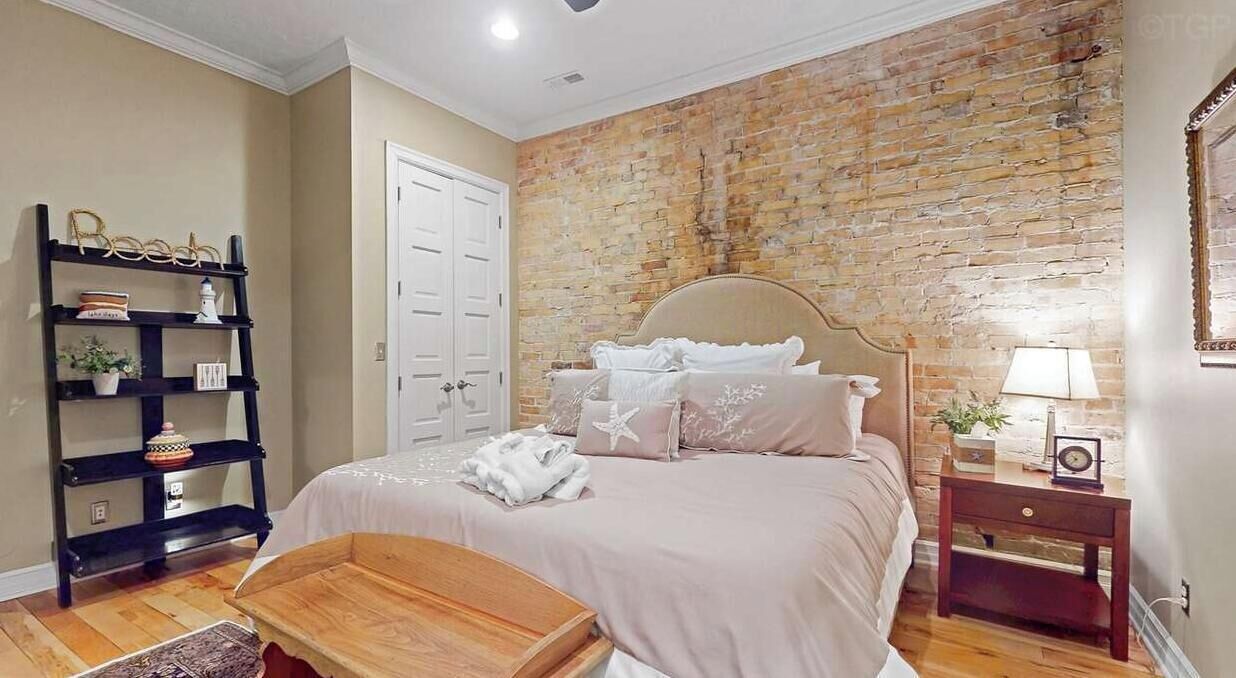 Una camera da letto con parete in mattoni, un letto con biancheria bianca, un comodino in legno, uno scaffale con oggetti decorativi e una porta bianca.