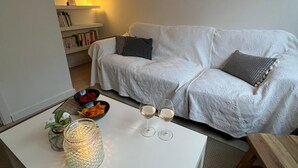Olohuoneessa on valkoinen sohva, lasinen kynttilälyhty, joka on asetettu sohvapöydälle, sekä kirjahylly.
