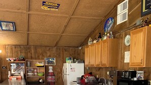 Cocina pequeña con armarios de madera, un refrigerador, un microondas y un lavamanos. Hay una mesa de comedor pequeña y varios utensilios de cocina en estantes.