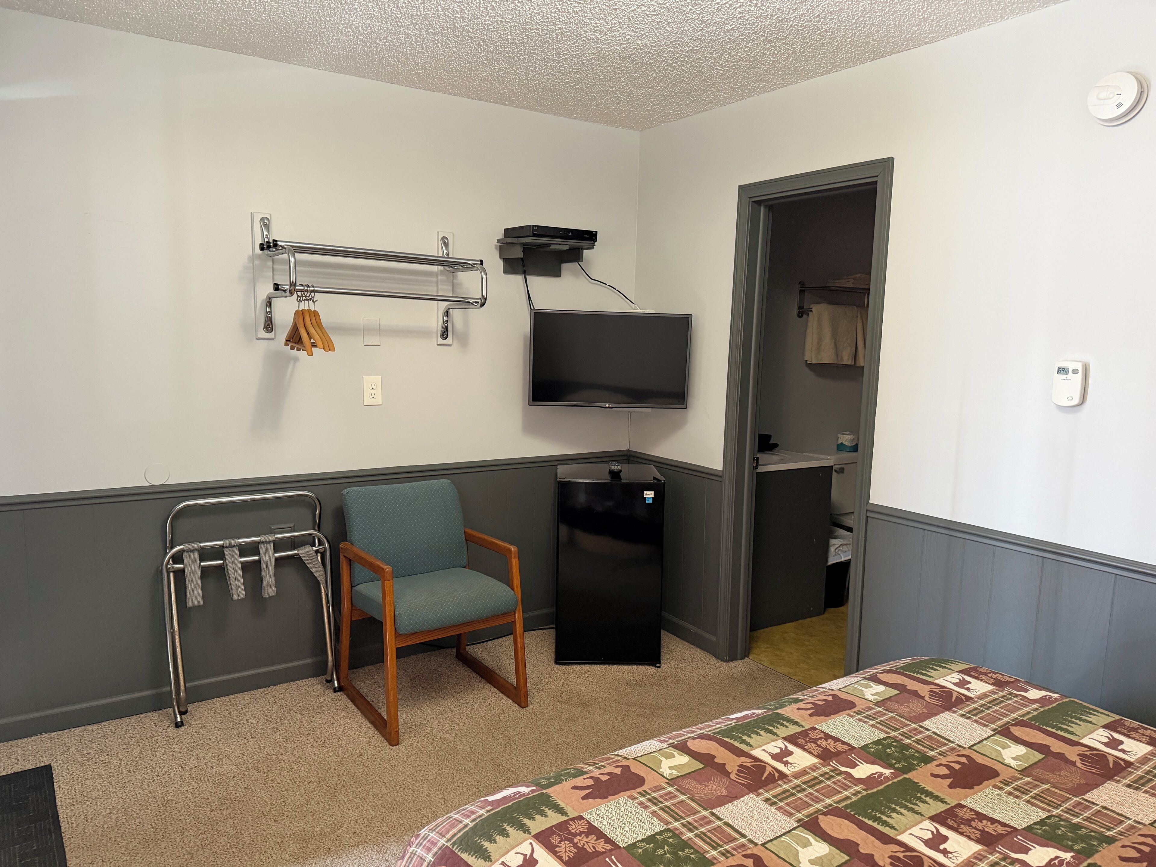Una habitación de hotel con cama, una silla, televisor, perchero y un escritorio pequeño.