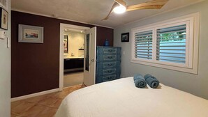 Una camera da letto con un letto, una ventola a soffitto, una finestra con persiane, un comò e un bagno visibile tramite una porta aperta.