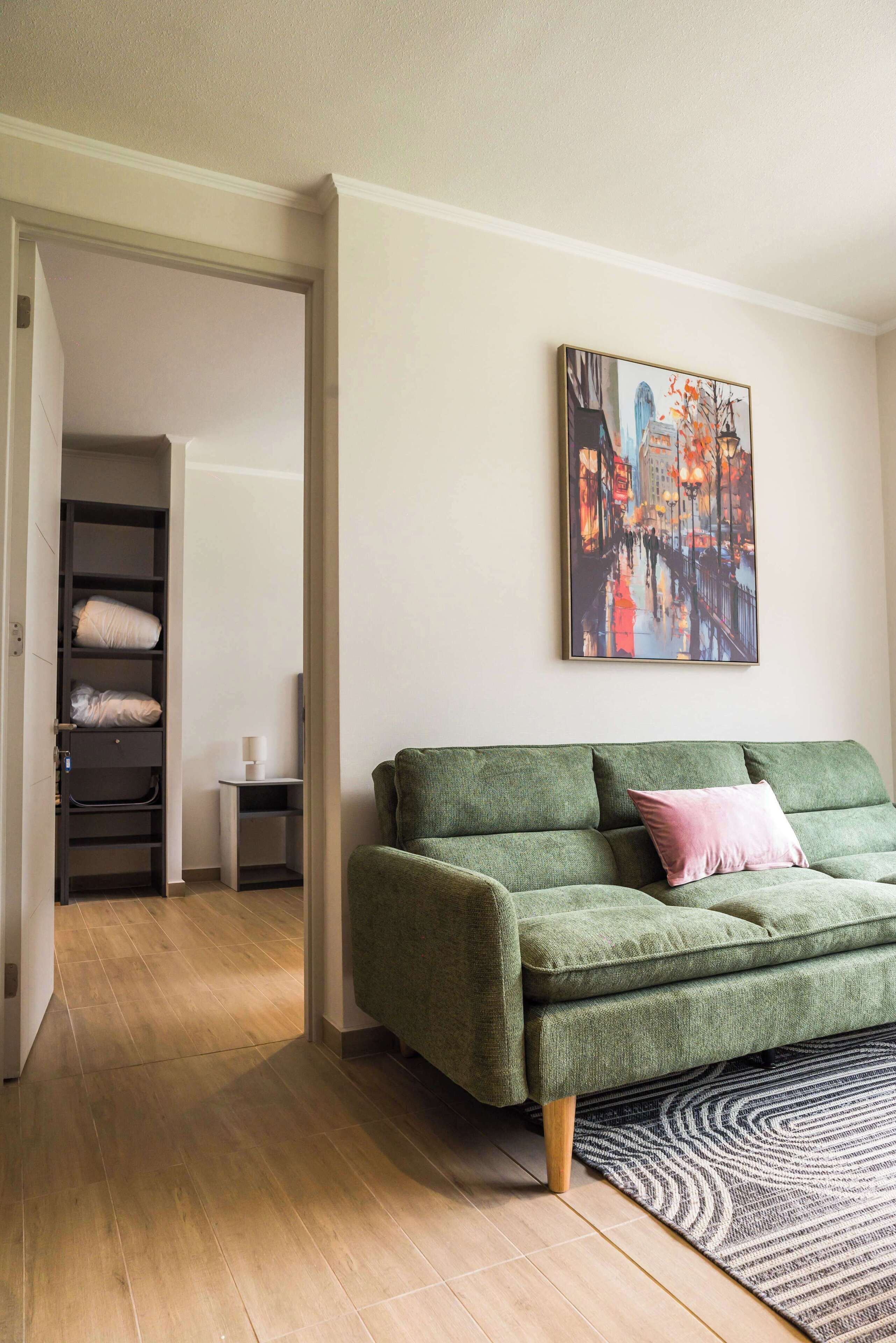 A green sofa with a pink pillow, a patterned rug, and a cityscape painting on the wall.