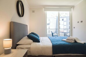 Une chambre d’hôtel avec un lit, une table de chevet avec une lampe, une fenêtre donnant sur des immeubles et un balcon.